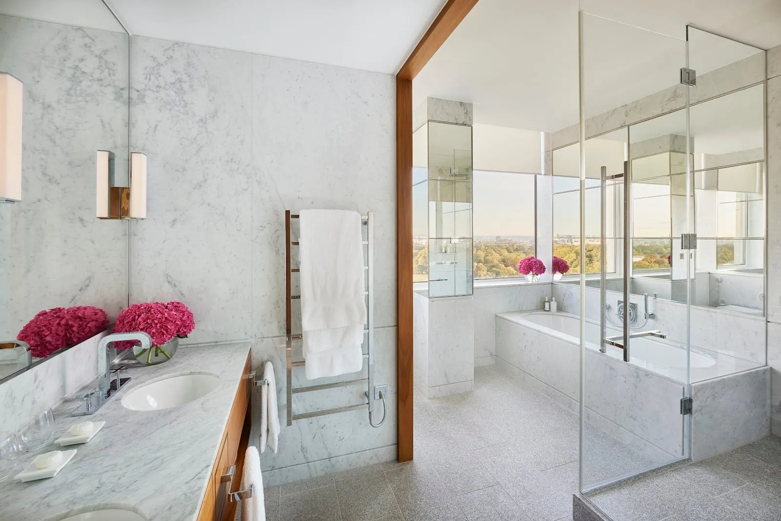 A suite bathroom at Royal Lancaster London in white marble, with twin basins in a walnut vanity, a bath and a glass shower enclosure beside a window looking down over the trees of the park