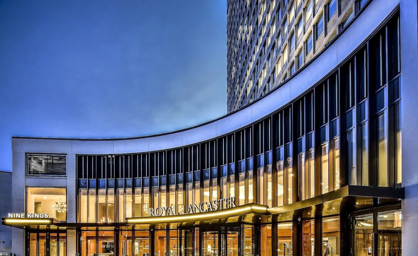 The curved glass and stone facade of Royal Lancaster London lit up at dusk, with the Royal Lancaster and Nine Kings signage over the entrance