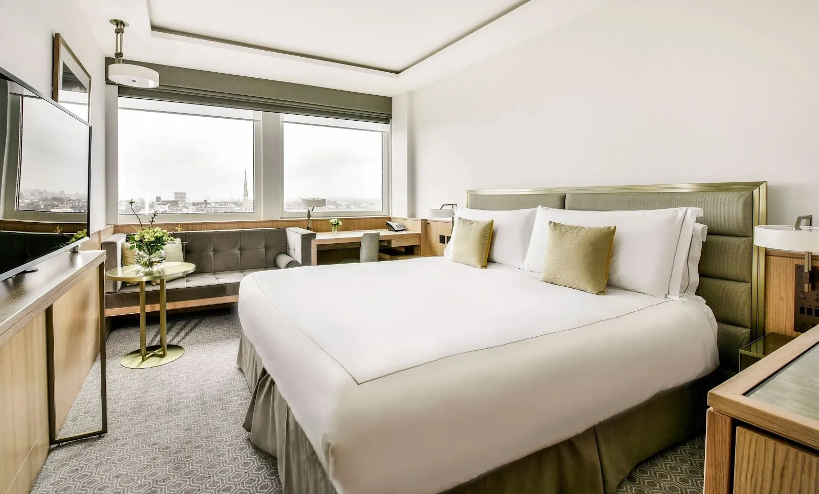 A double room at Royal Lancaster London with a wide bed, a green and brass padded headboard, a grey velvet sofa and a desk under a long window looking over rooftops and a church spire