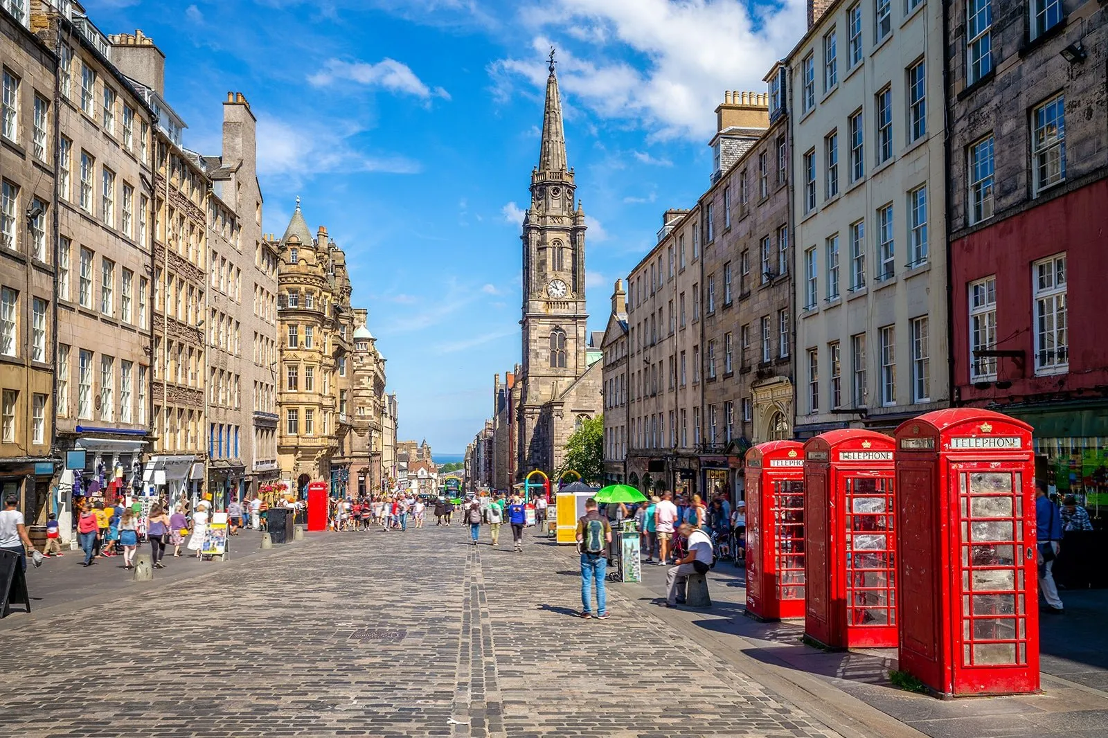 Cobbled Royal Mile lined with tenement buildings, red telephone boxes and a church spire, with crowds of visitors walking its length