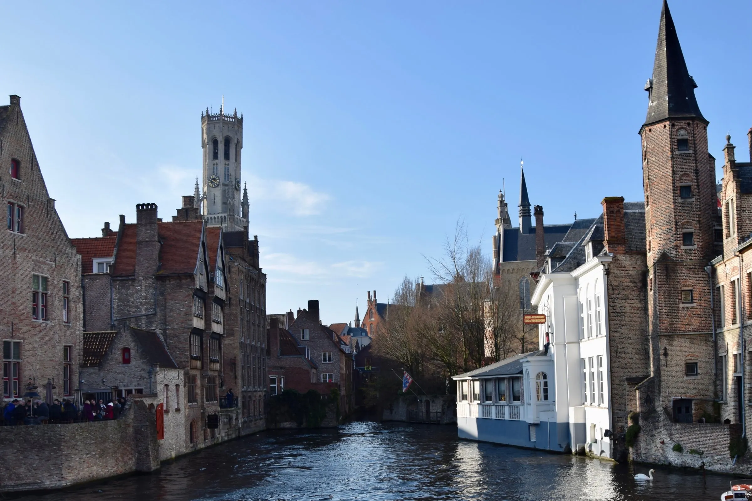 The Rozenhoedkaai canal in Bruges on a clear day, with the Belfry's tower rising above stepped gable rooftops and a white gabled house at the water's edge