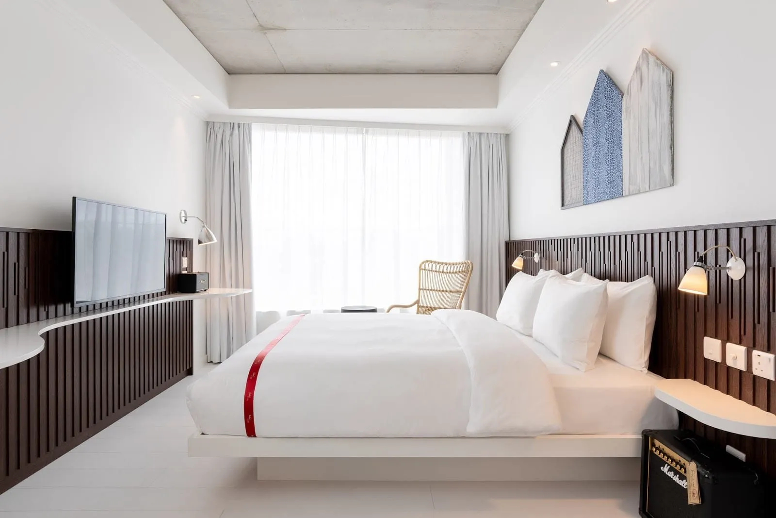 A room at Ruby Zoe with a white platform bed crossed by a red ribbon, dark slatted wood panelling behind the bed and TV, a rattan armchair by a wide curtained window and a Marshall guitar amp on the floor