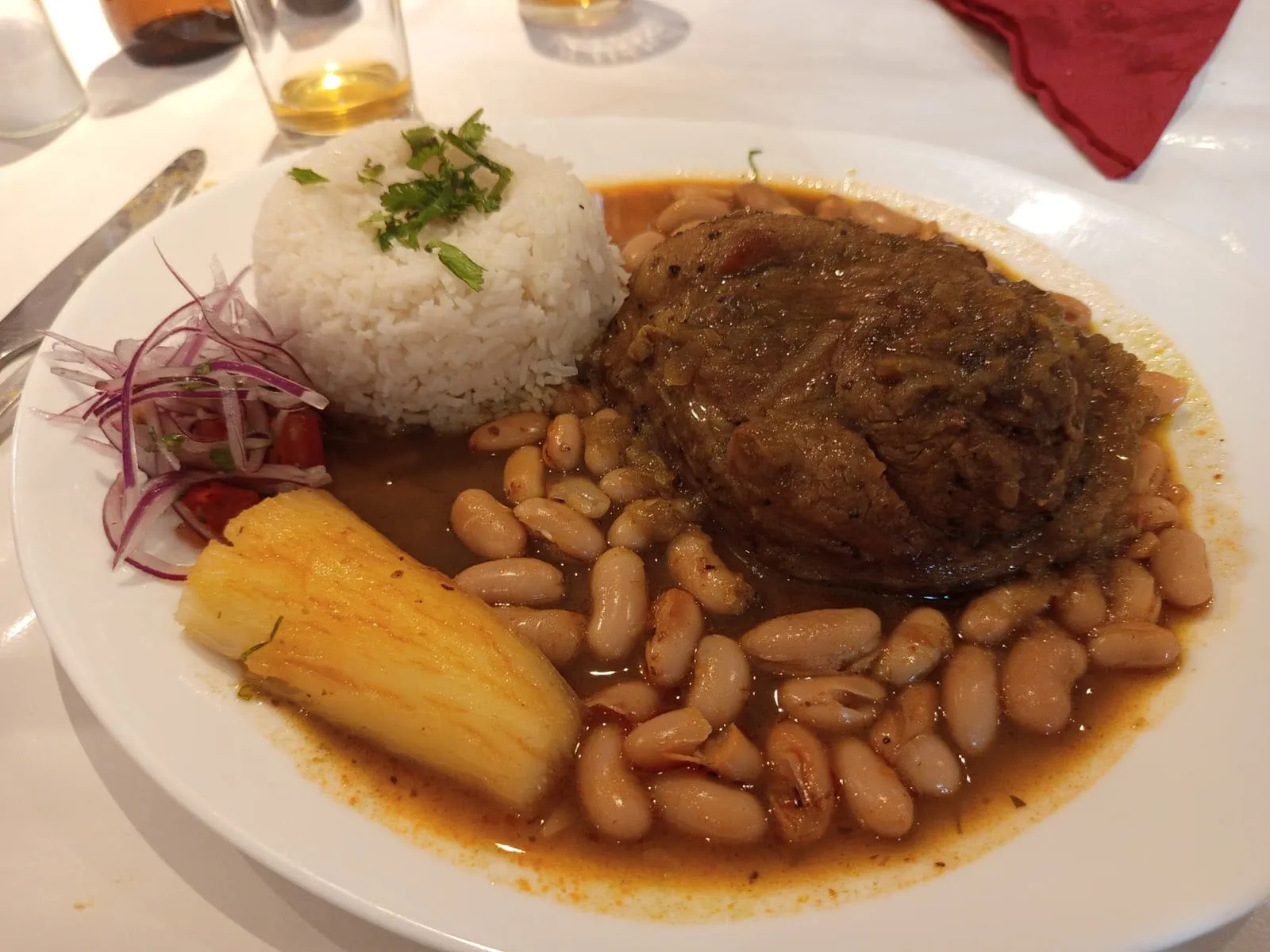 A white plate on a white tablecloth holding beef slow-cooked in a dark coriander sauce with canary beans, a moulded dome of white rice, a length of boiled cassava and a pile of sliced red onion
