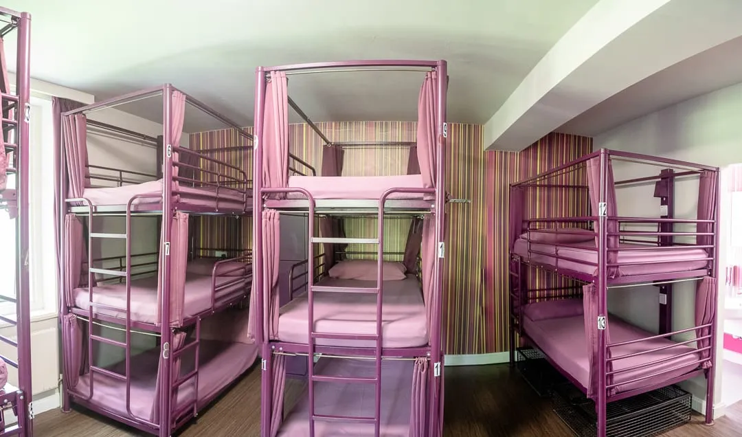 A dorm room at Safestay London Kensington Holland Park, with curtained pod-style bunks and pink-and-green striped feature wallpaper