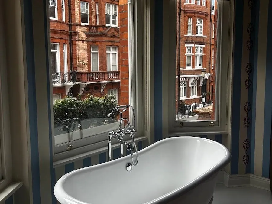 A roll-top bath with a chrome hand shower set in the window of a room at San Domenico House, with blue-and-cream striped walls and red-brick mansion blocks across the street