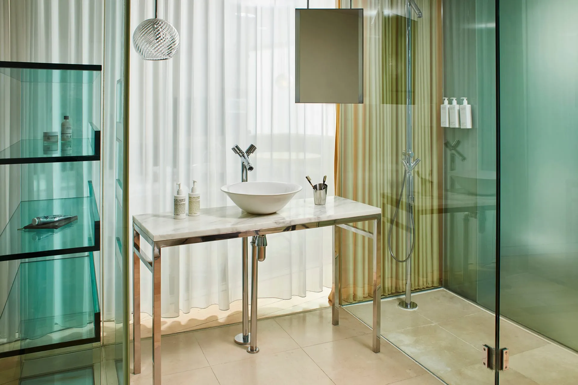 The bathroom at Sanderson London, with a white vessel basin on a marble console table, sheer curtains and a glass-walled shower