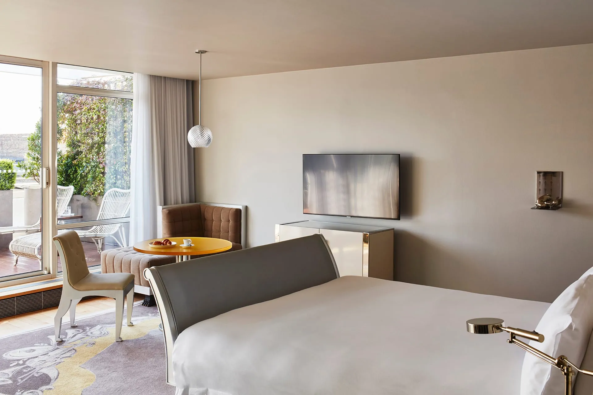 A Sanderson London room with a grey sleigh bed, a round yellow breakfast table set with coffee and a croissant, and a private terrace beyond the glass doors