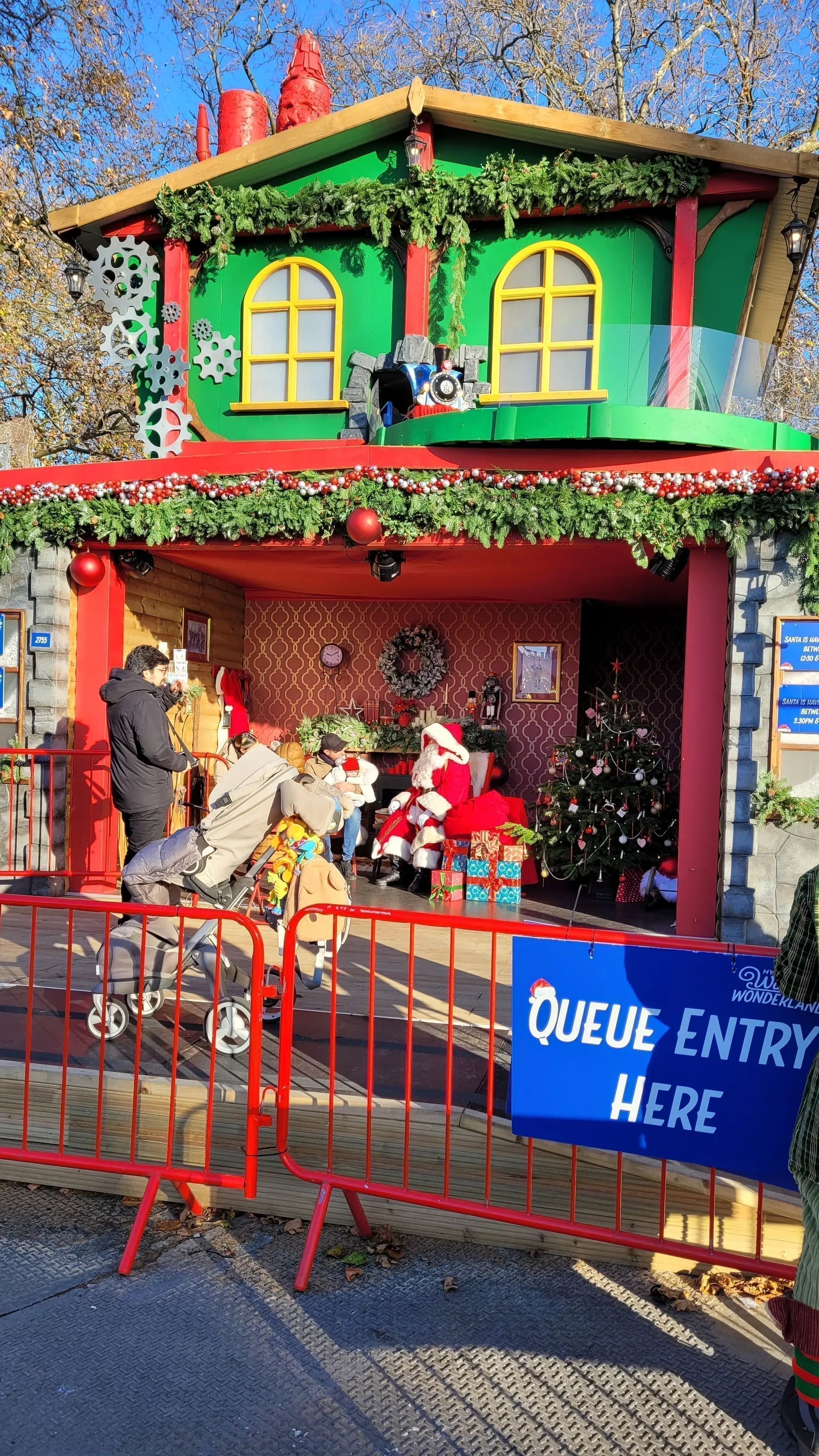 Inside the green and red Santa's Grotto hut at Winter Wonderland, with a queue sign, a decorated Christmas tree and Father Christmas visible