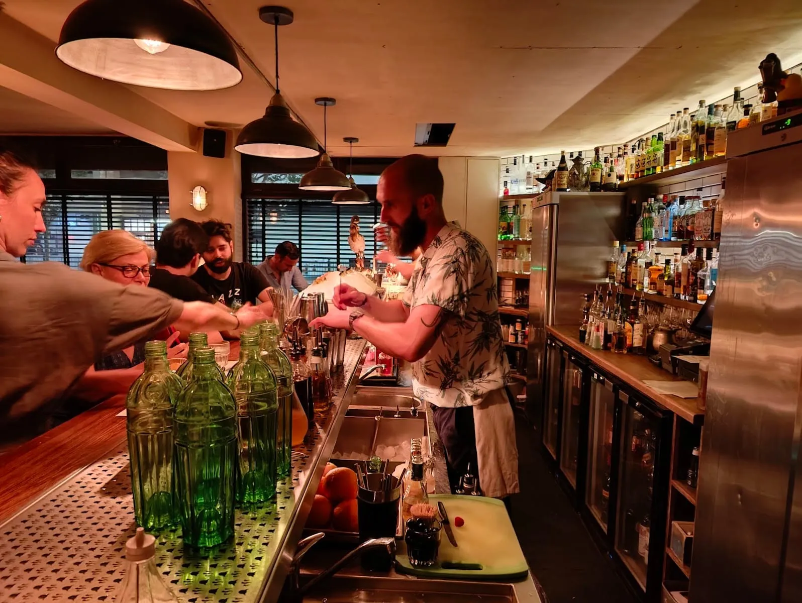 A bartender making drinks behind the bar, with customers seated along the counter, green glass bottles on the rail and shelves of spirits behind him