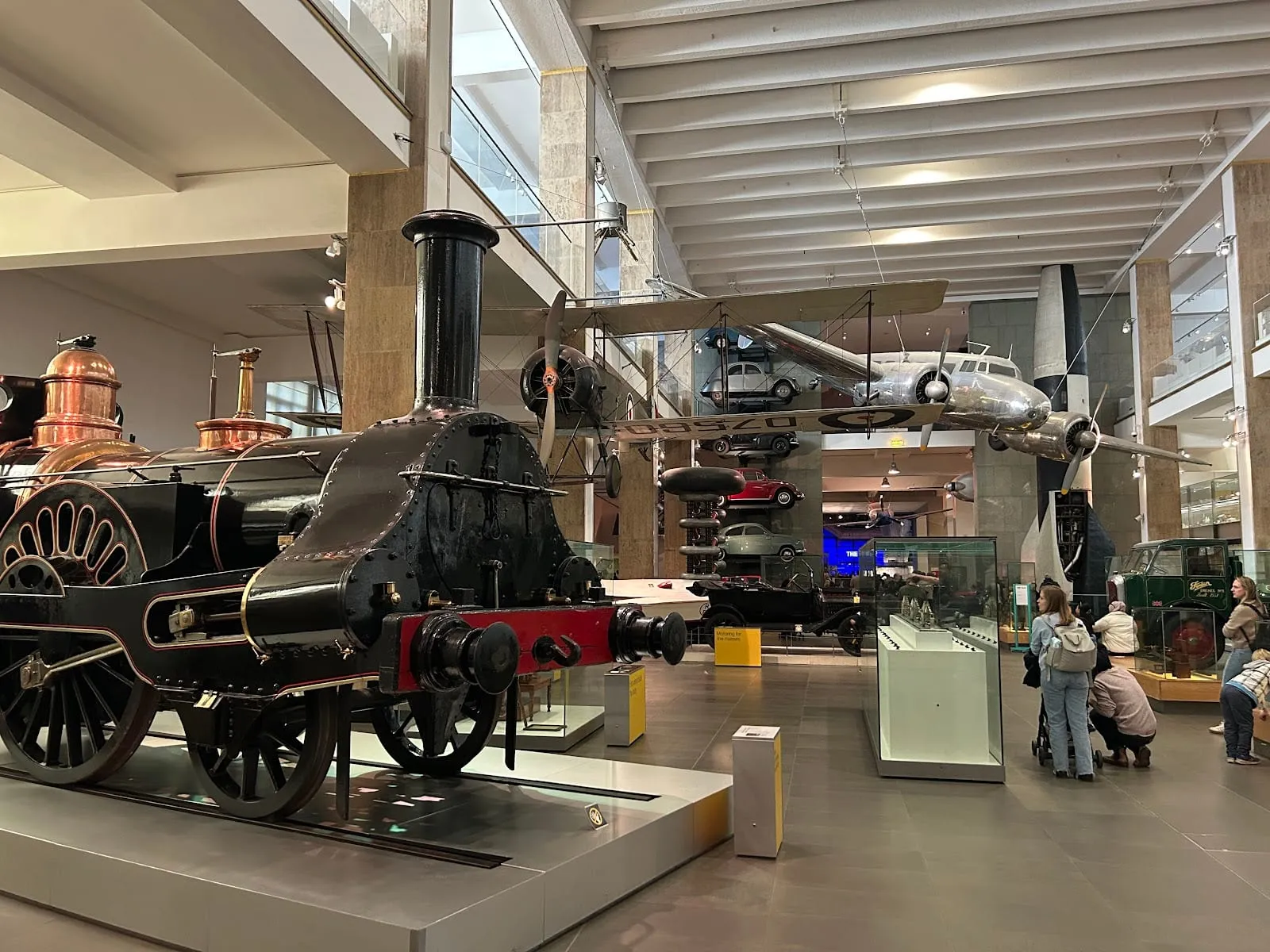 A black Victorian steam locomotive in the foreground with vintage cars and a biplane suspended overhead in the Science Museum's main hall