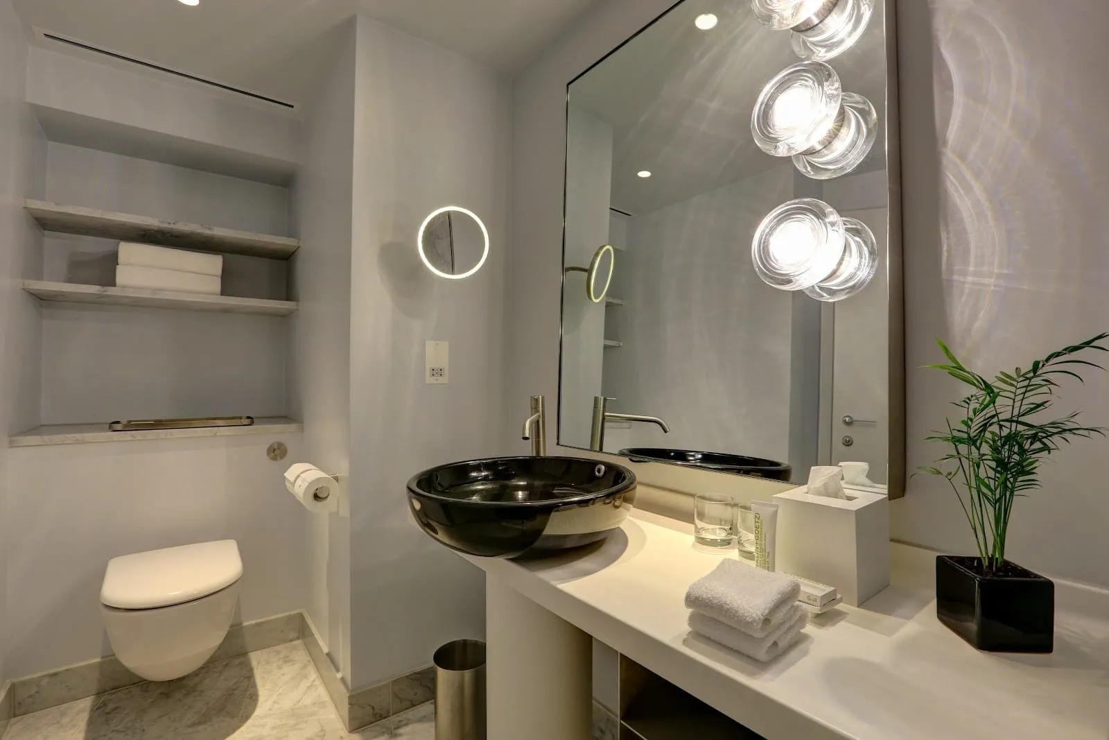 A bathroom at Sea Containers London with a black bowl basin on a white counter, a mirror lit by large glass bulbs and a wall-hung toilet