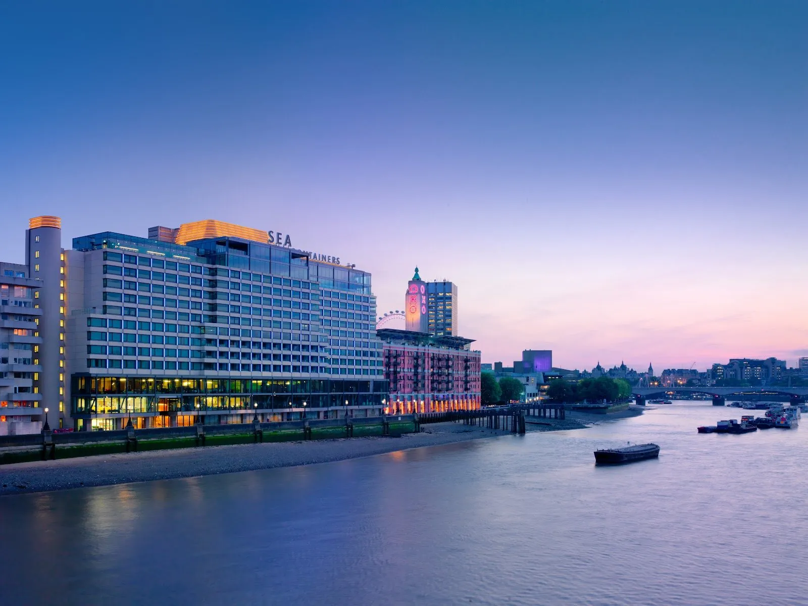 Sea Containers London lit up at dusk on the South Bank riverside path, with the OXO Tower and passing barges on the Thames beside it