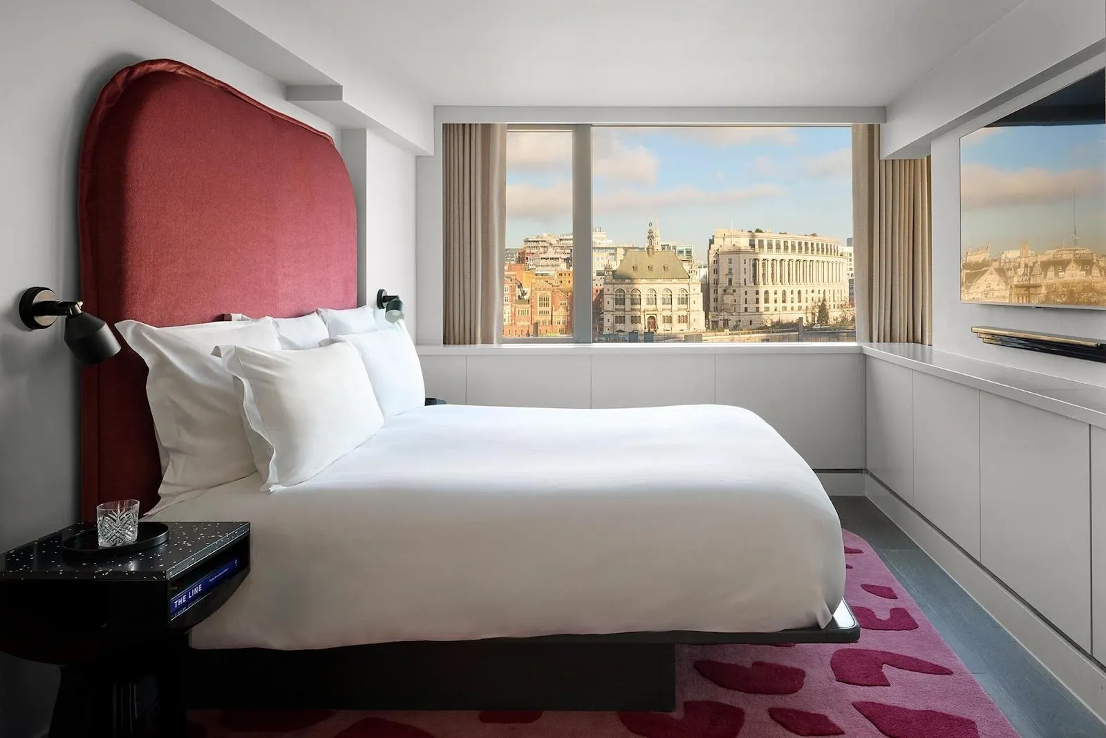 A room at Sea Containers London with a curved wine-red headboard and a patterned pink rug, looking out over the City of London skyline