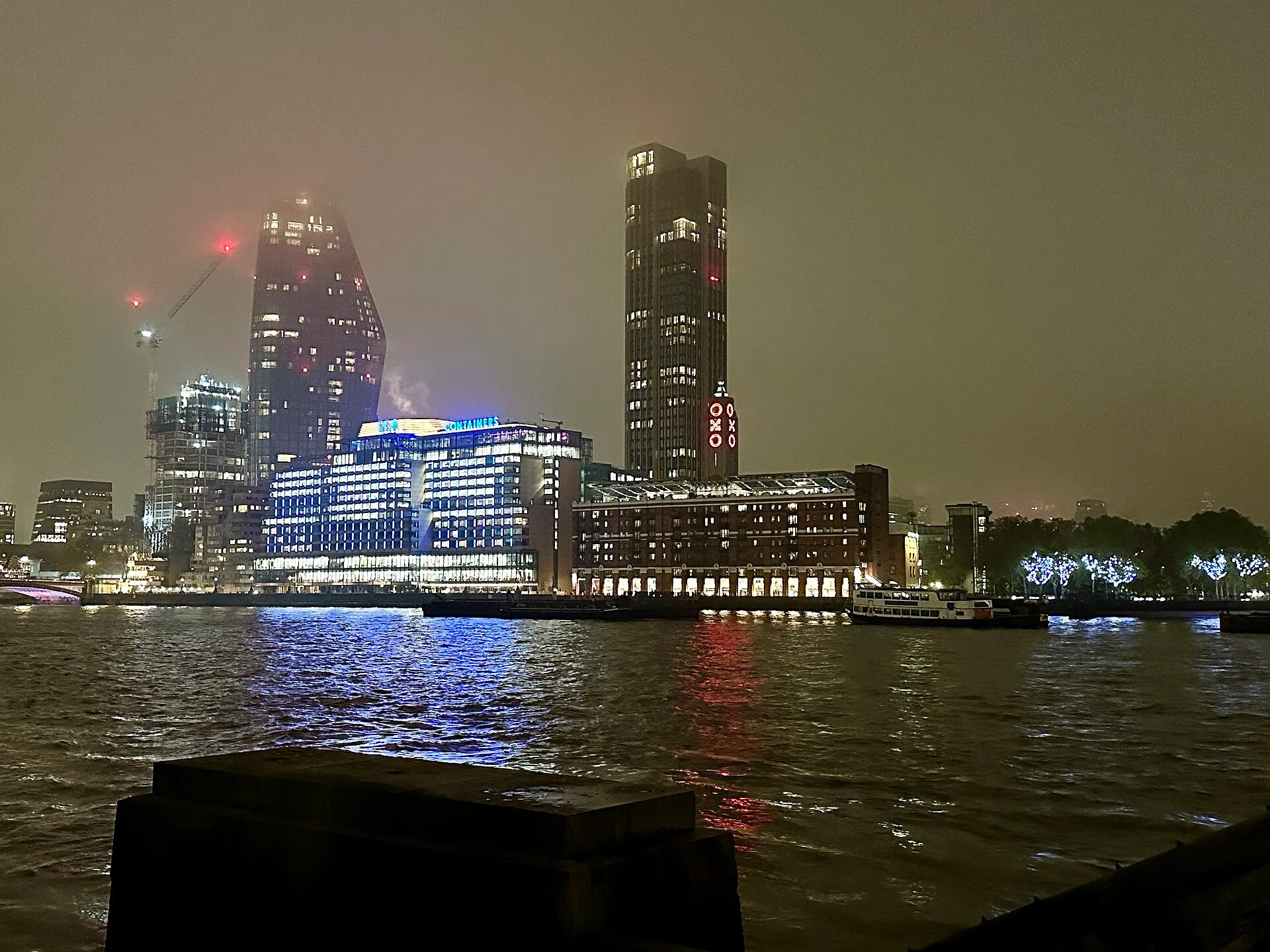 The South Bank at night from across the Thames: Sea Containers lit blue with its name on the roof, the red OXO Tower sign beside it, and the curved One Blackfriars tower in low cloud behind, with a river boat moored in front