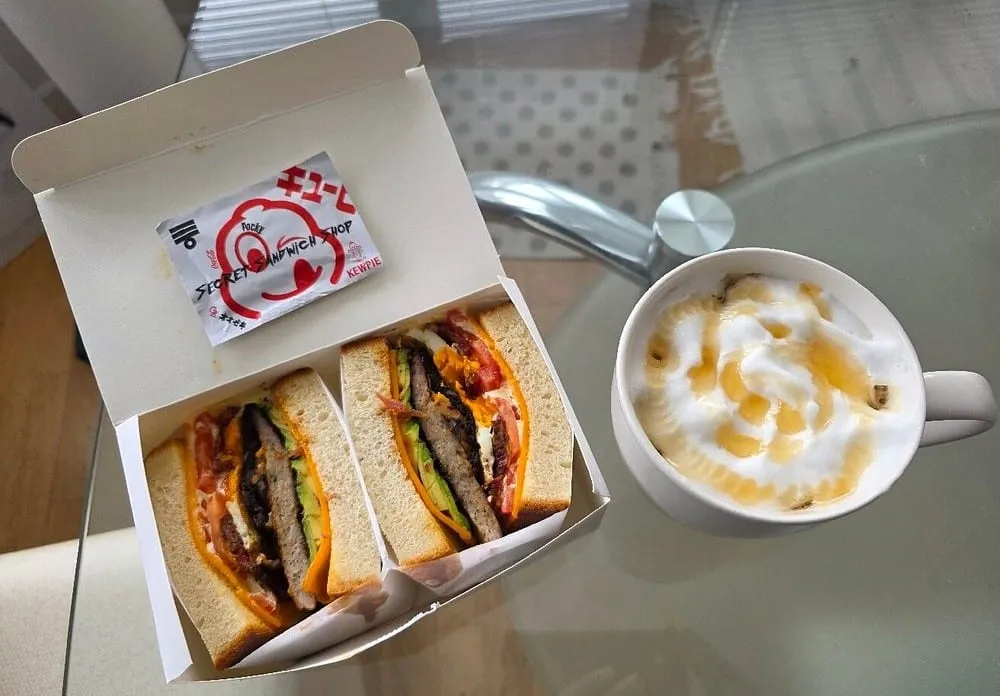 A sando cut in two in a white takeaway box, crustless white bread around a filling of patty, avocado, tomato and egg, with a branded Kewpie sachet tucked in beside it