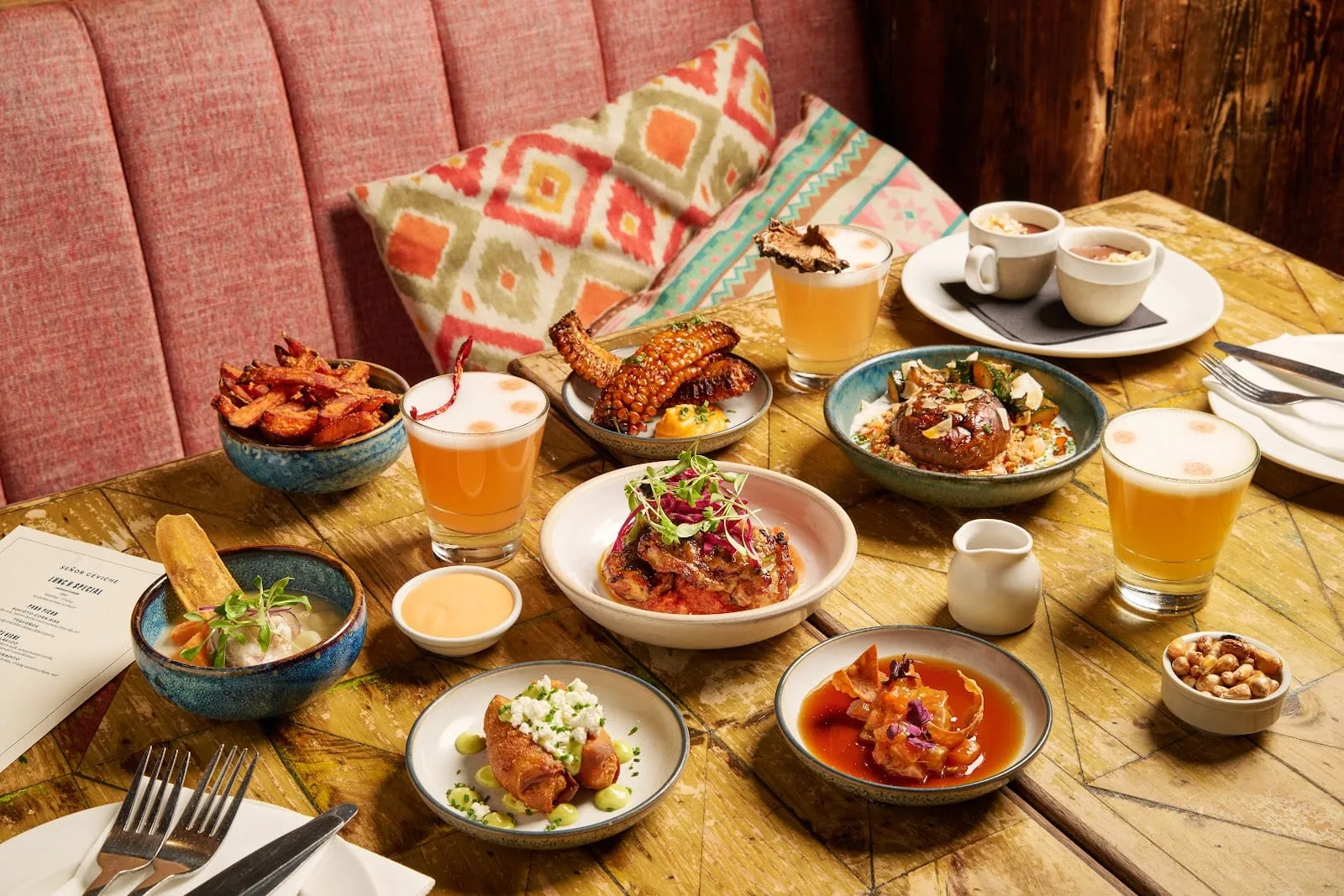 A table of Peruvian small plates — sweet potato fries, charred corn, a bowl of ceviche, fried tequeños, salmon tiradito and three foam-topped pisco sours — on a painted wooden table against a pink banquette, with a menu card reading Señor Ceviche Lunes Special