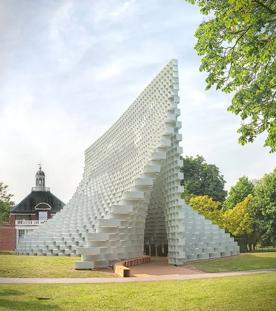 A pointed white lattice-block pavilion in Kensington Gardens, tapering from a tall peak down to ground level, with a period building visible behind it