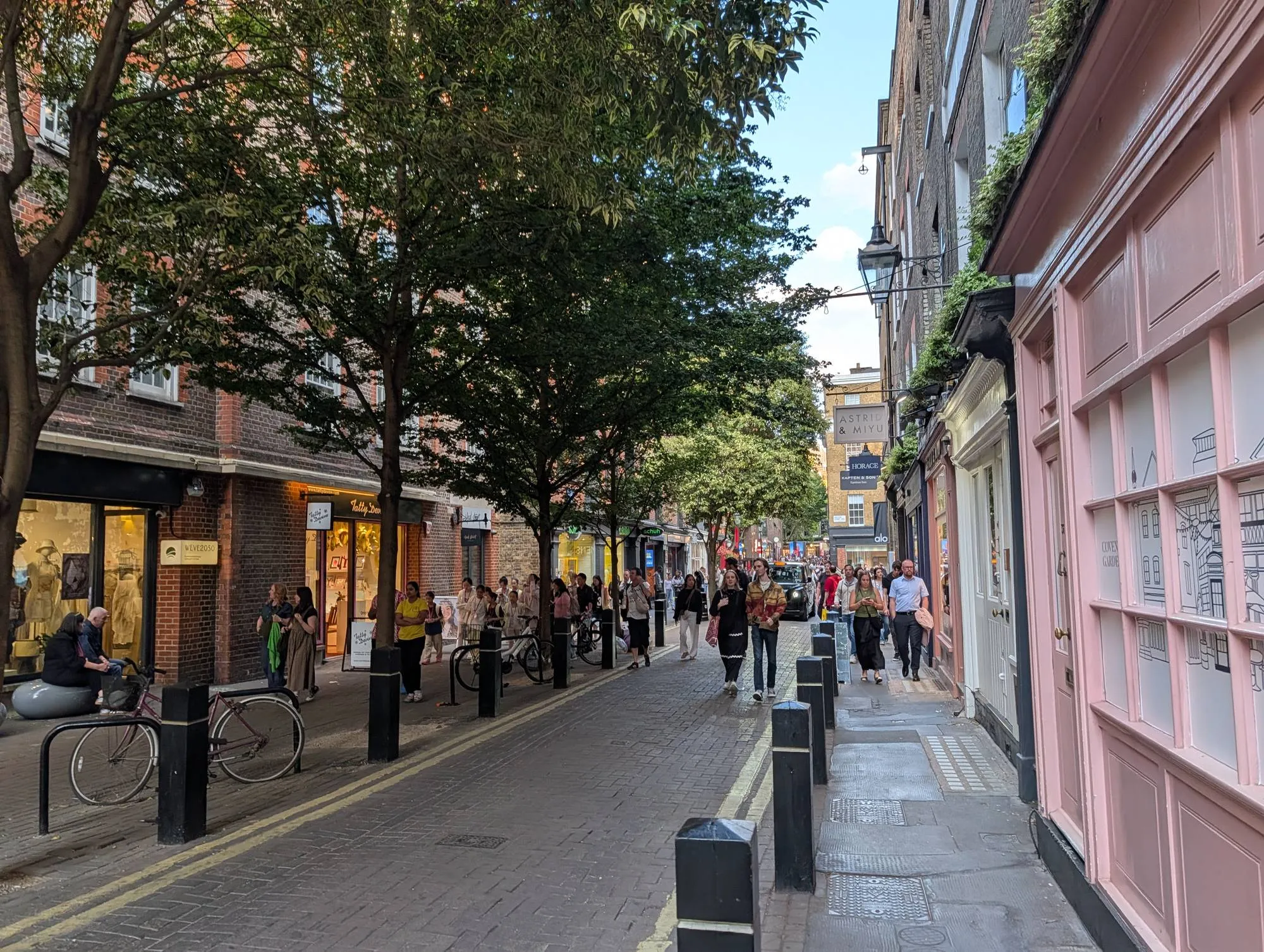 Shoppers on a tree-lined pedestrian street in Seven Dials, with small brand shopfronts on both sides