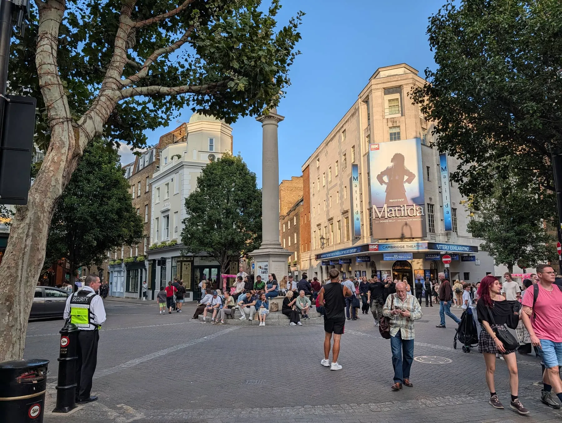 The sundial pillar at the Seven Dials junction, with the Cambridge Theatre behind it and people sitting on the steps