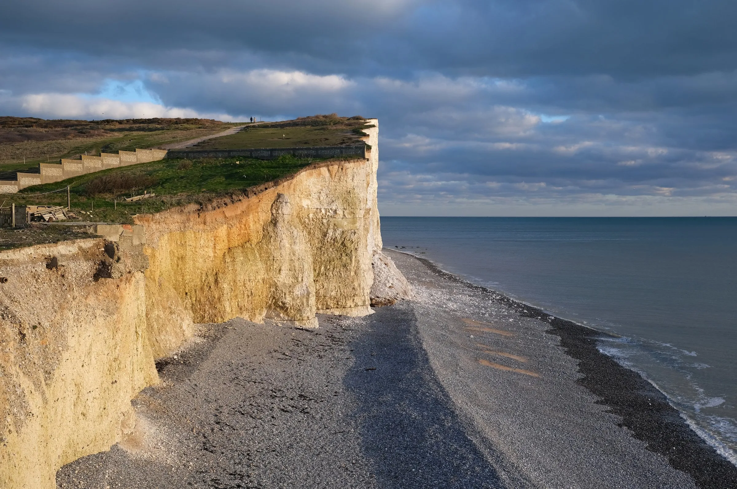 The chalk cliffs of the Seven Sisters seen from Birling Gap at golden hour, with two walkers on the clifftop path and a wartime pillbox base in the foreground