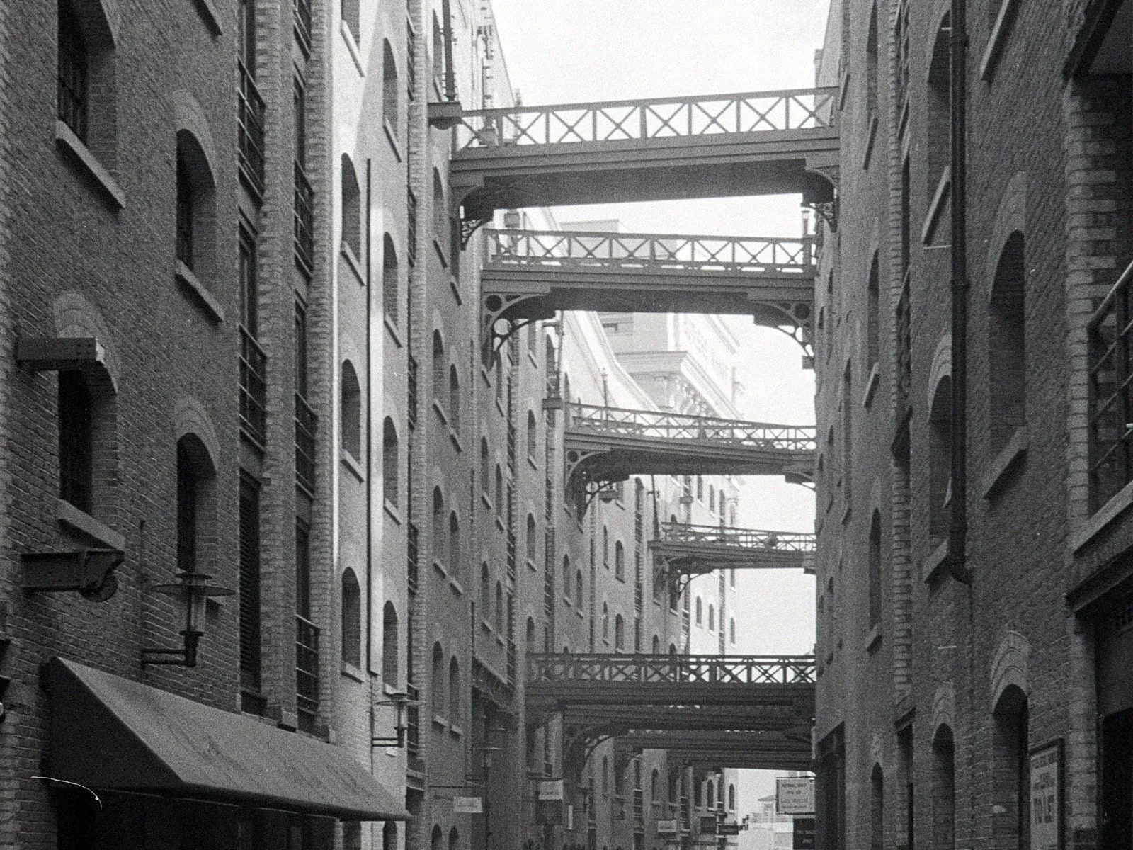 Shad Thames in black and white: tall Victorian brick warehouses either side of a narrow street, crossed overhead by a row of iron gantry walkways receding into the distance