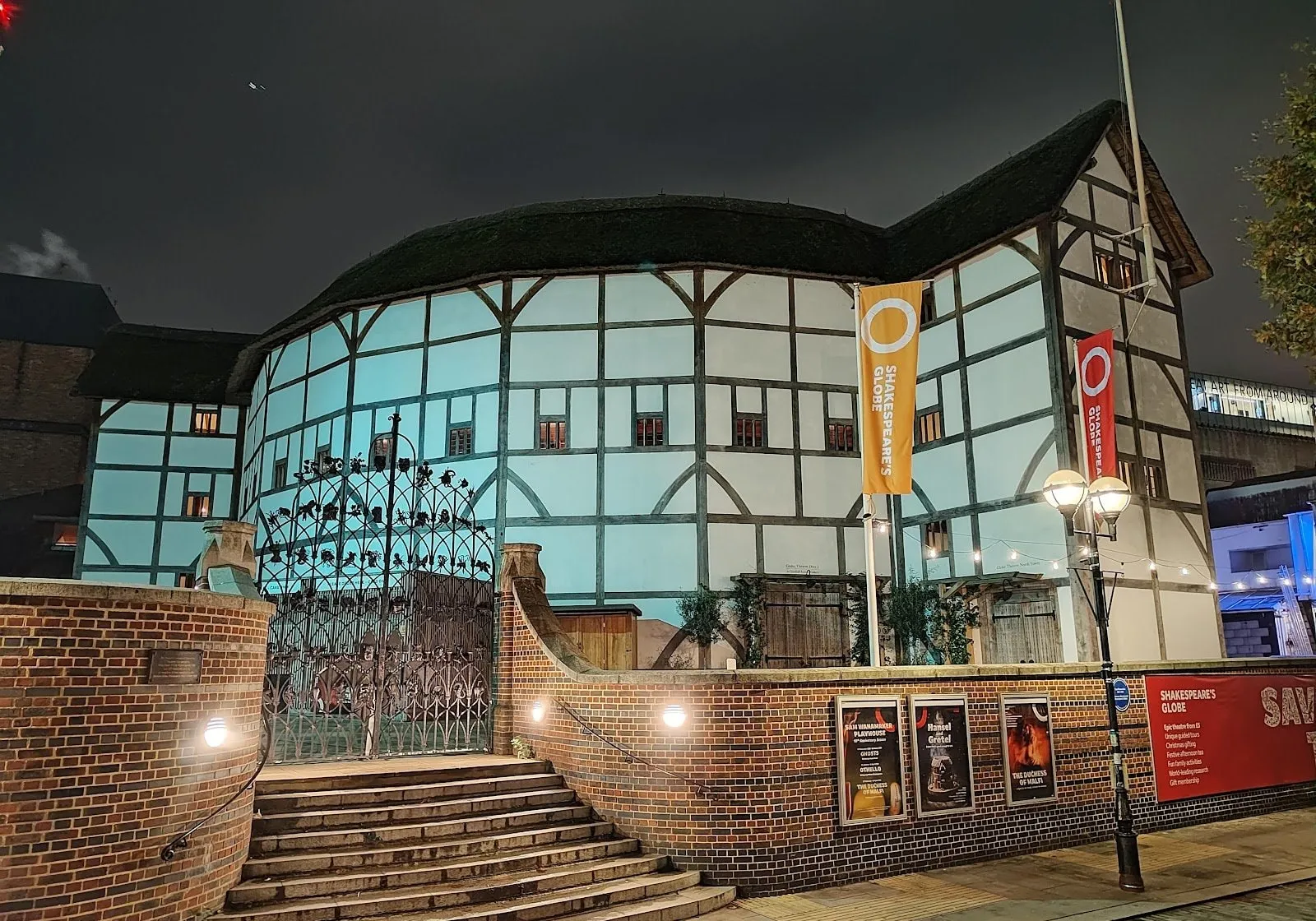 Shakespeare's Globe at night, its white-plastered timber frame and thatched roof lit up behind the ironwork gates and steps on the riverside, with Globe banners and show posters on the brick wall