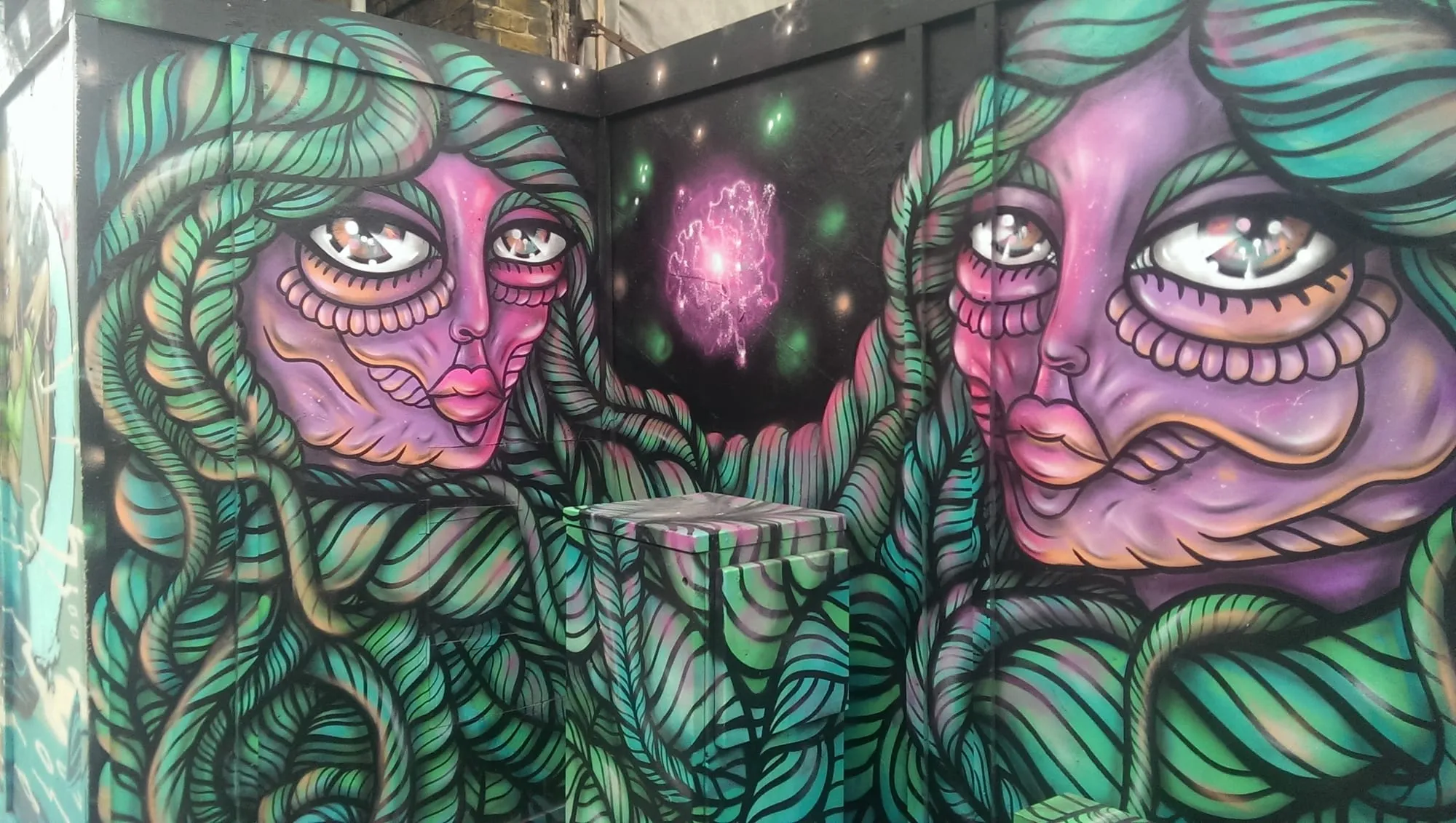 Two large mural faces with green rope-like hair and a pink nebula painted between them, on a Shoreditch corner