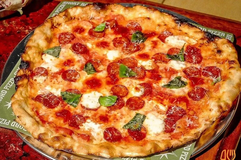 A Short Road pizza on a metal tray, the rim puffed and charred in patches, topped with cupped pepperoni, torn mozzarella, grated parmesan and fresh basil, sitting on green Short Road Pizza paper