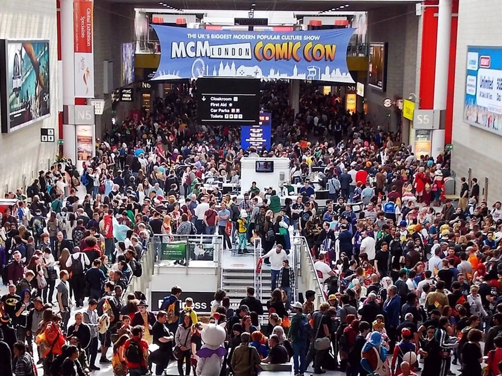 Crowds filling the concourse under an MCM London Comic Con banner, with signage pointing to the cloakroom, first aid, car parks and toilets