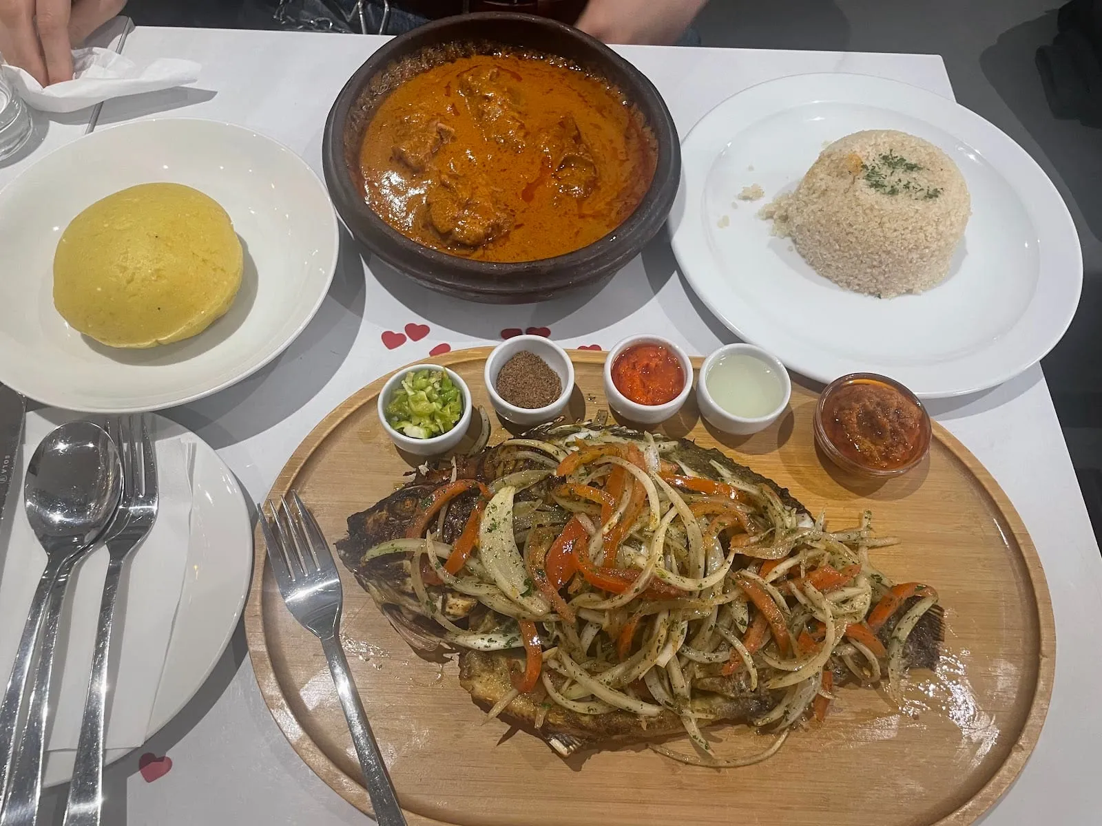An Ivorian table from above: a whole grilled fish under a heap of sliced onion and sweet pepper on a wooden board, a moulded dome of pale granular attiéké on one plate, an earthenware pot of orange-red stew, and a row of small pots of condiments including chopped green chilli, ground brown spice and red chilli sauce