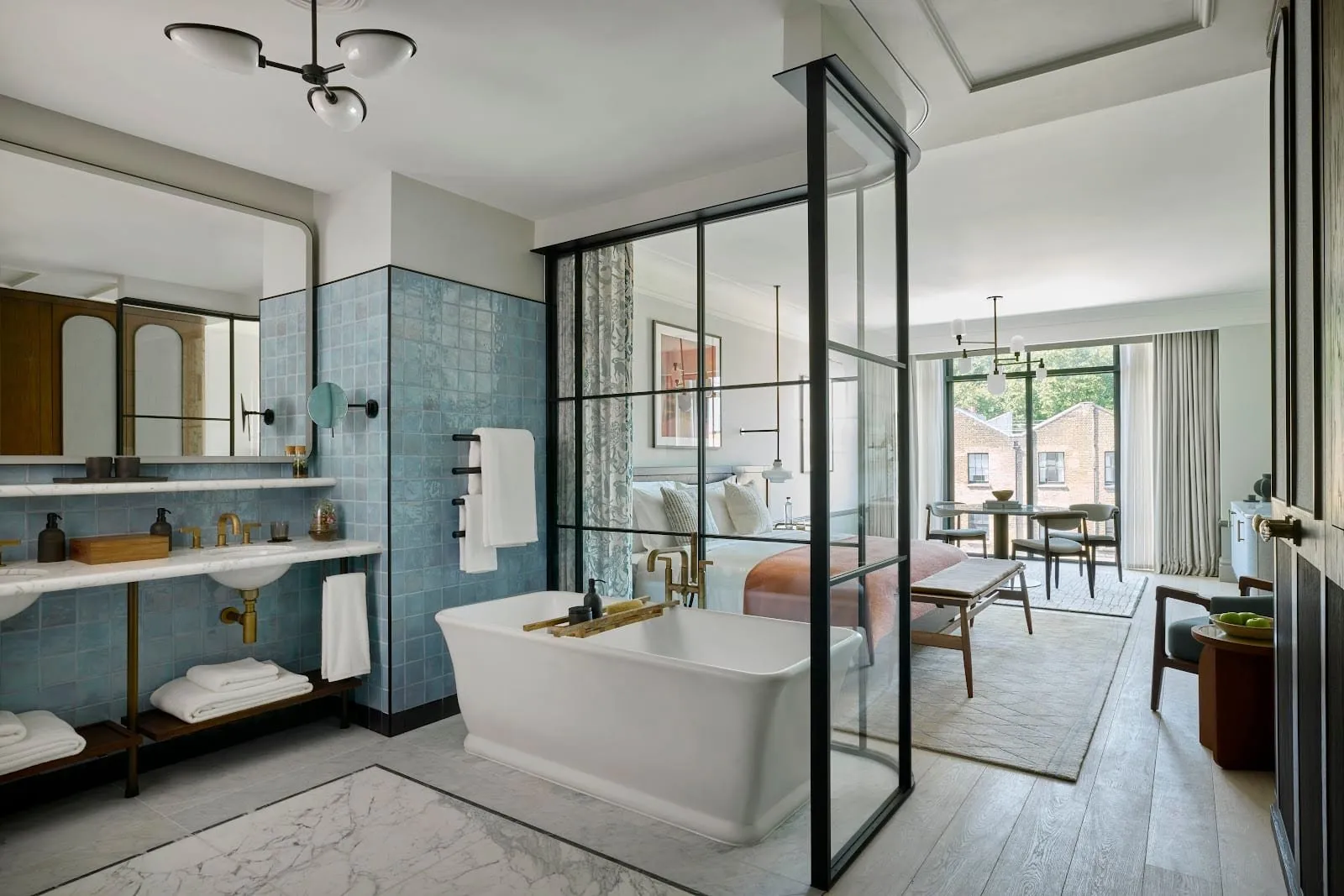 A larger room at Six Senses London where a freestanding white bath stands behind a black-framed glass screen, with twin basins on a marble shelf against blue tiles and the bed and a dining table beyond