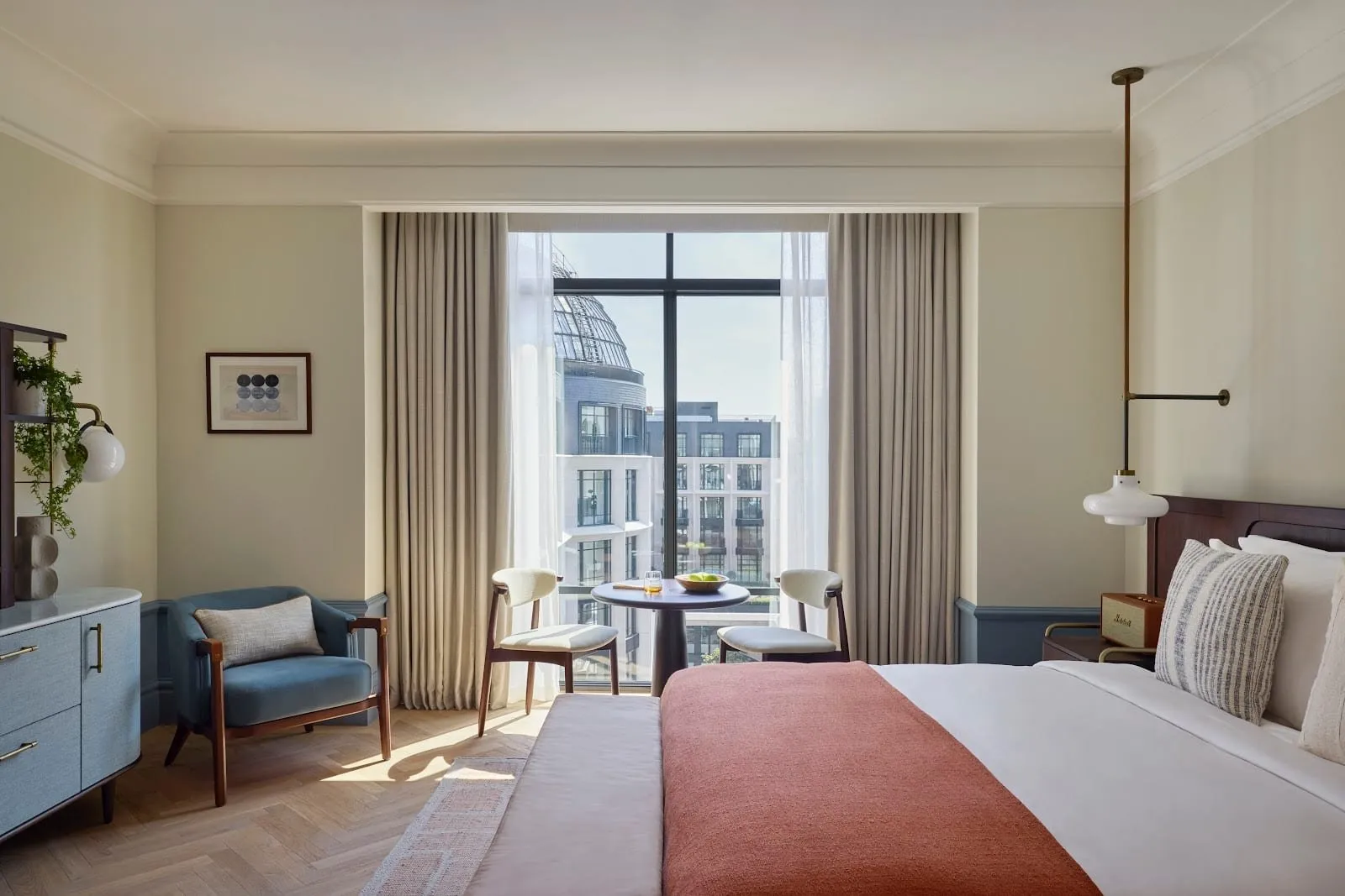 A room at Six Senses London with cream walls, a herringbone oak floor, a rust-coloured throw on the bed, a blue armchair and a table for two at a floor-to-ceiling window looking across to the glass dome of The Whiteley