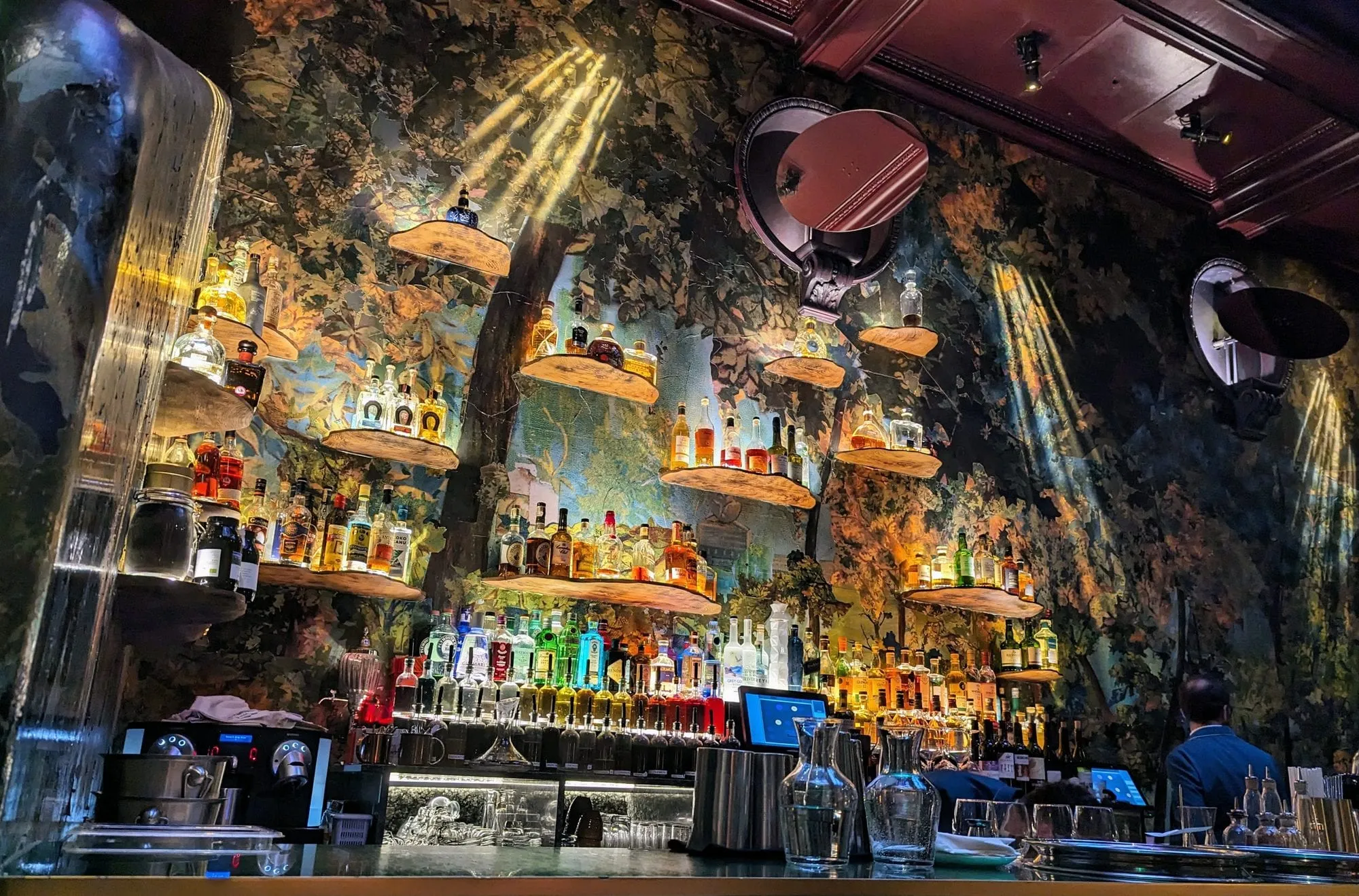 The bar at Sketch, a woodland mural lit in shafts of light, with bottles on floating wooden shelves under a dark red panelled ceiling