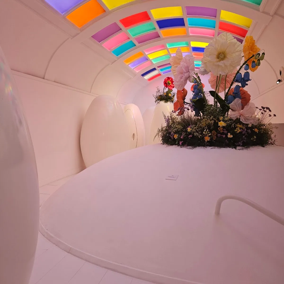 One of sketch's white egg-shaped lavatory pods, beneath a ceiling painted in bands of colour, next to a display of oversized paper flowers
