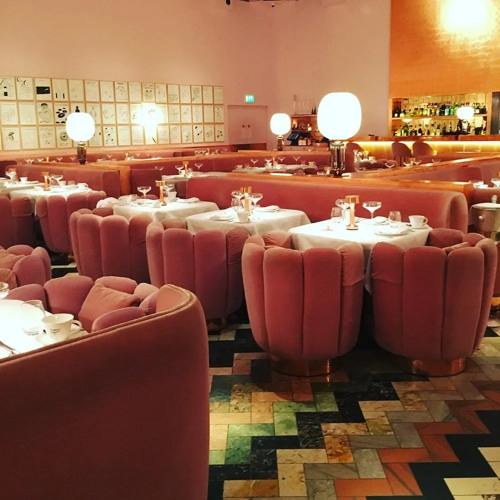 The Gallery at Sketch before its 2022 rebuild: pink velvet scallop-backed chairs around white-clothed tables, David Shrigley's framed drawings covering the wall, on a harlequin marble floor