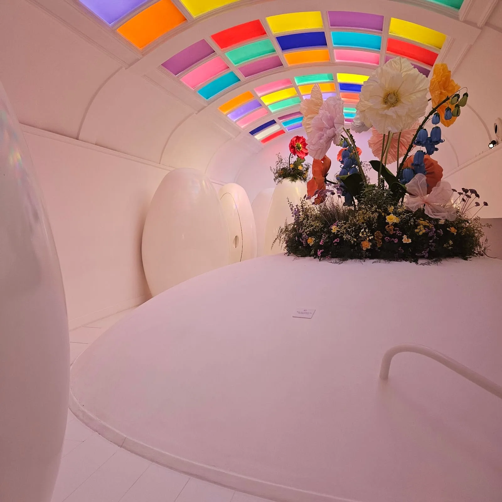 The white egg-shaped lavatory pods at Sketch under a ceiling of coloured glass panels, with a large paper-flower installation between them