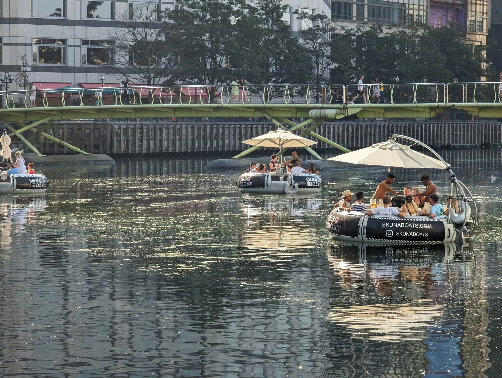 Small round boats with sun umbrellas and built-in barbecues on the dock at Canary Wharf, with people cooking on board