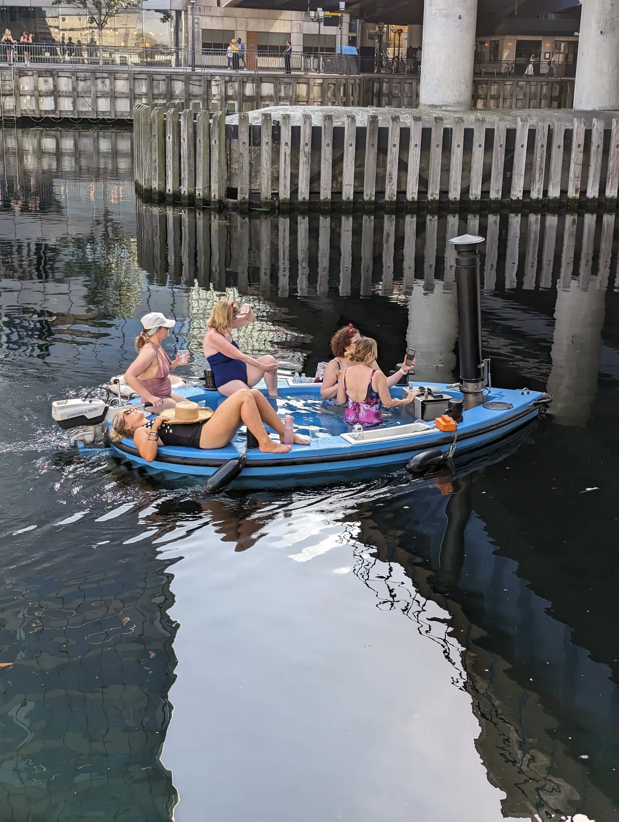 People sitting in a wood-fired hot tub built into a small boat on the dock at Canary Wharf