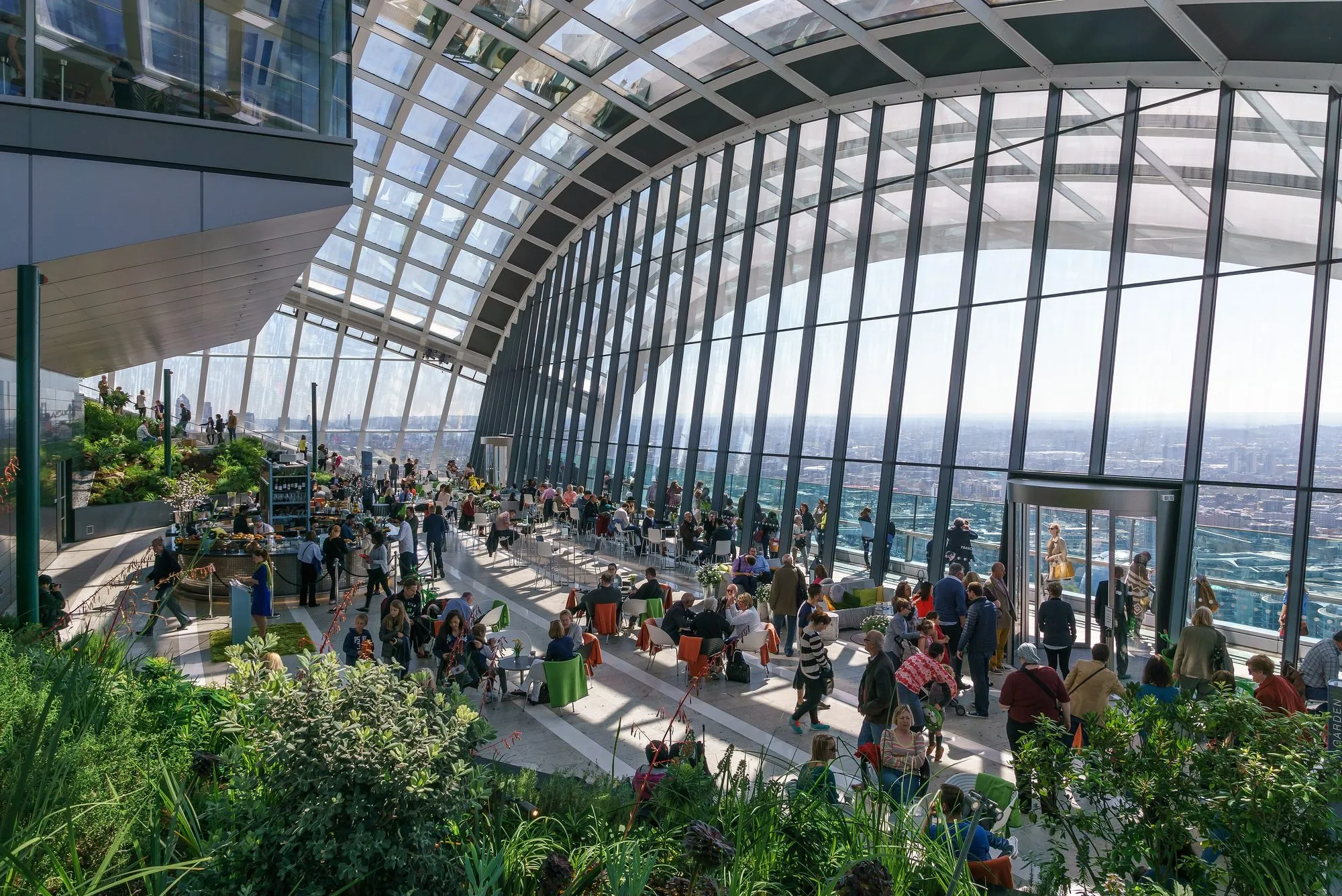 The tall curved glass roof of the Sky Garden over a busy café floor, with planted terraces on the left and visitors at the windows