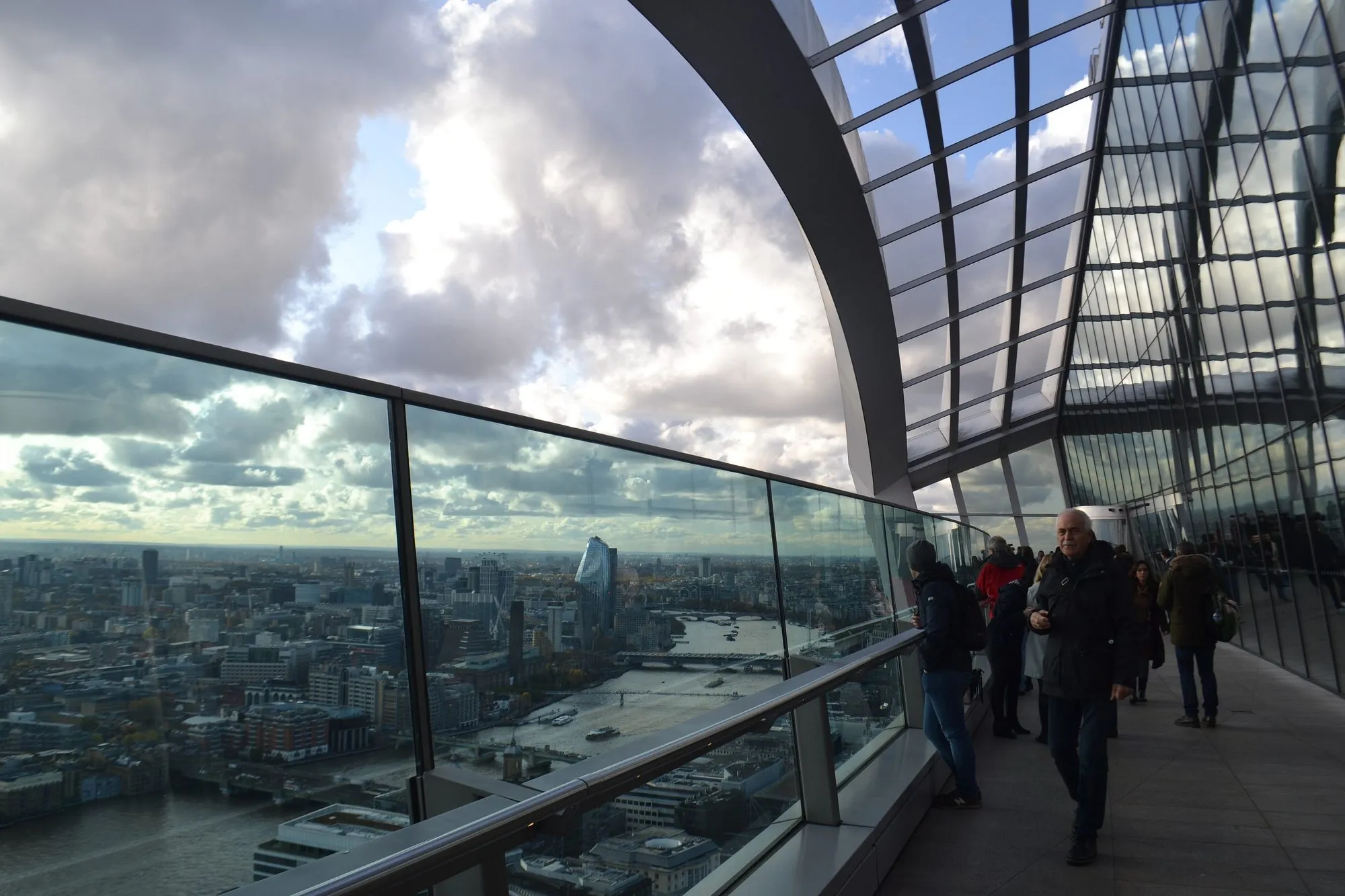 Visitors on the open-air terrace behind a glass balustrade, the Thames and the towers of Blackfriars below a cloudy sky