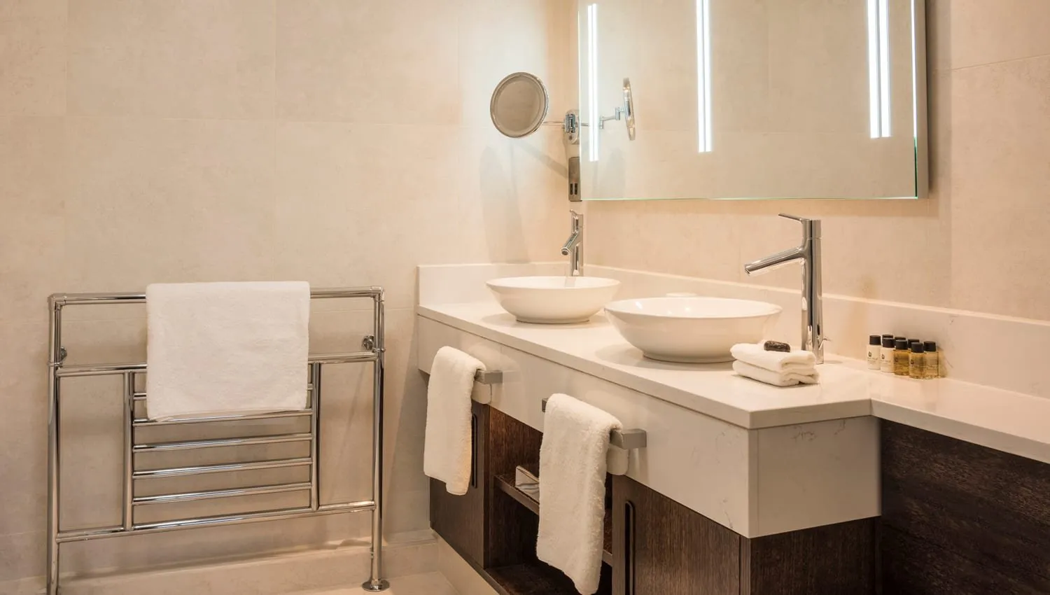 A bathroom at Sloane Place with two white bowl basins on a white stone counter over a dark wood vanity, a lit mirror and a chrome towel rack