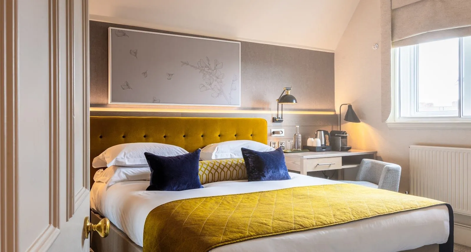 A room at Sloane Place under a sloping ceiling, with a mustard velvet headboard, navy velvet cushions, a mustard quilted runner, a desk with a kettle and coffee machine, and a small window