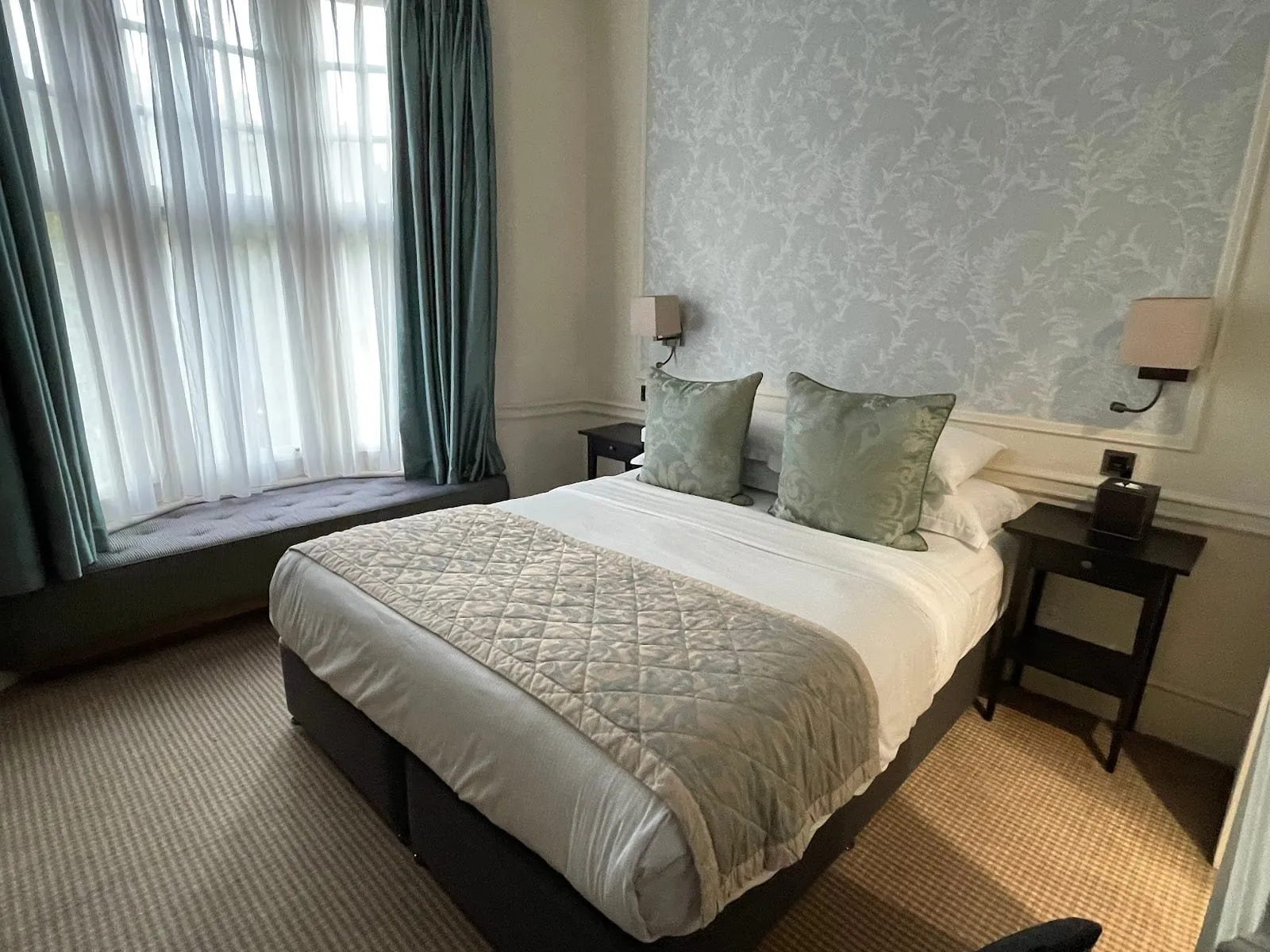 A room at the Sloane Square Hotel with a double bed, a pale blue floral panel behind it, dark wood bedside tables, and a padded window seat under a tall window hung with sheer and teal curtains