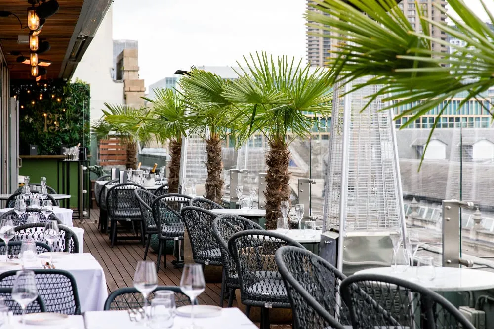 The rooftop terrace at Smiths of Smithfield: white-clothed tables and black woven armchairs on decking, potted fan palms along a glass balustrade, tall gas patio heaters, and Smithfield rooftops beyond