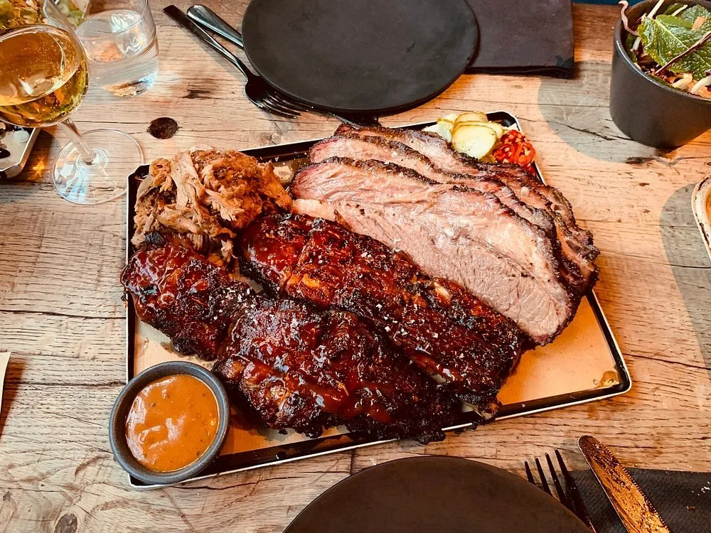 A tray of sliced beef brisket with a black bark and a pink smoke ring, glazed pork ribs and pulled pork, with pickles and a pot of sauce on a wooden table