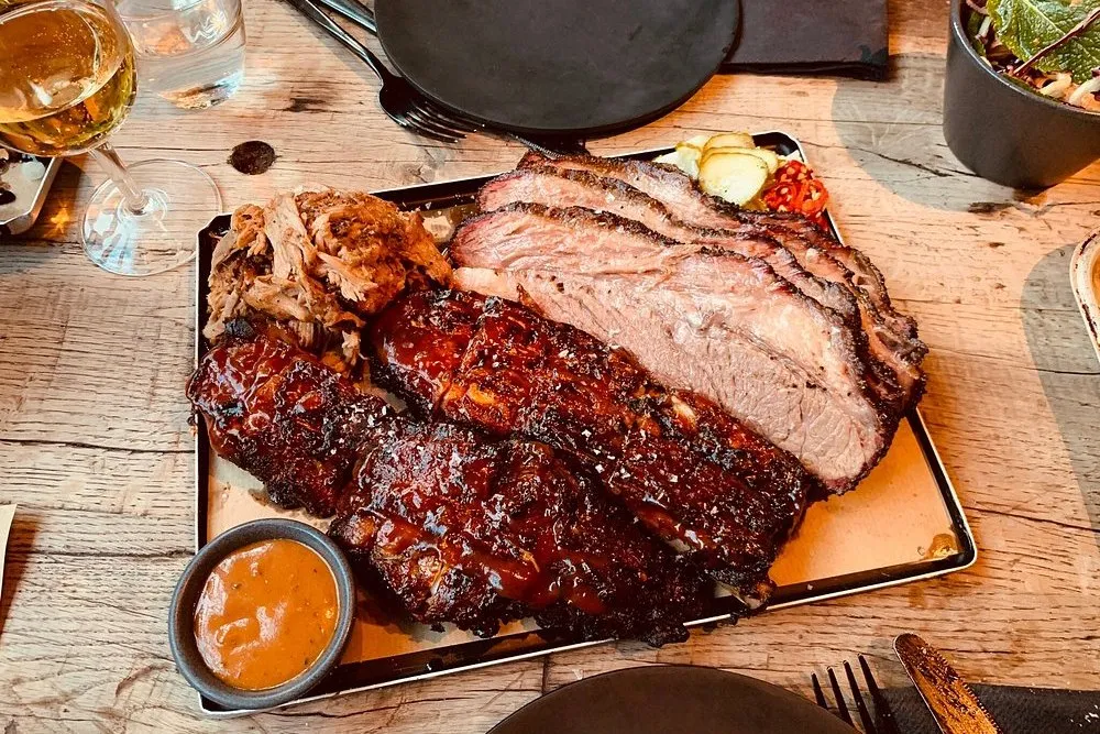 A tray of sliced beef brisket with a black bark and a pink smoke ring, glazed pork ribs and pulled pork, with pickles and a pot of sauce on a wooden table