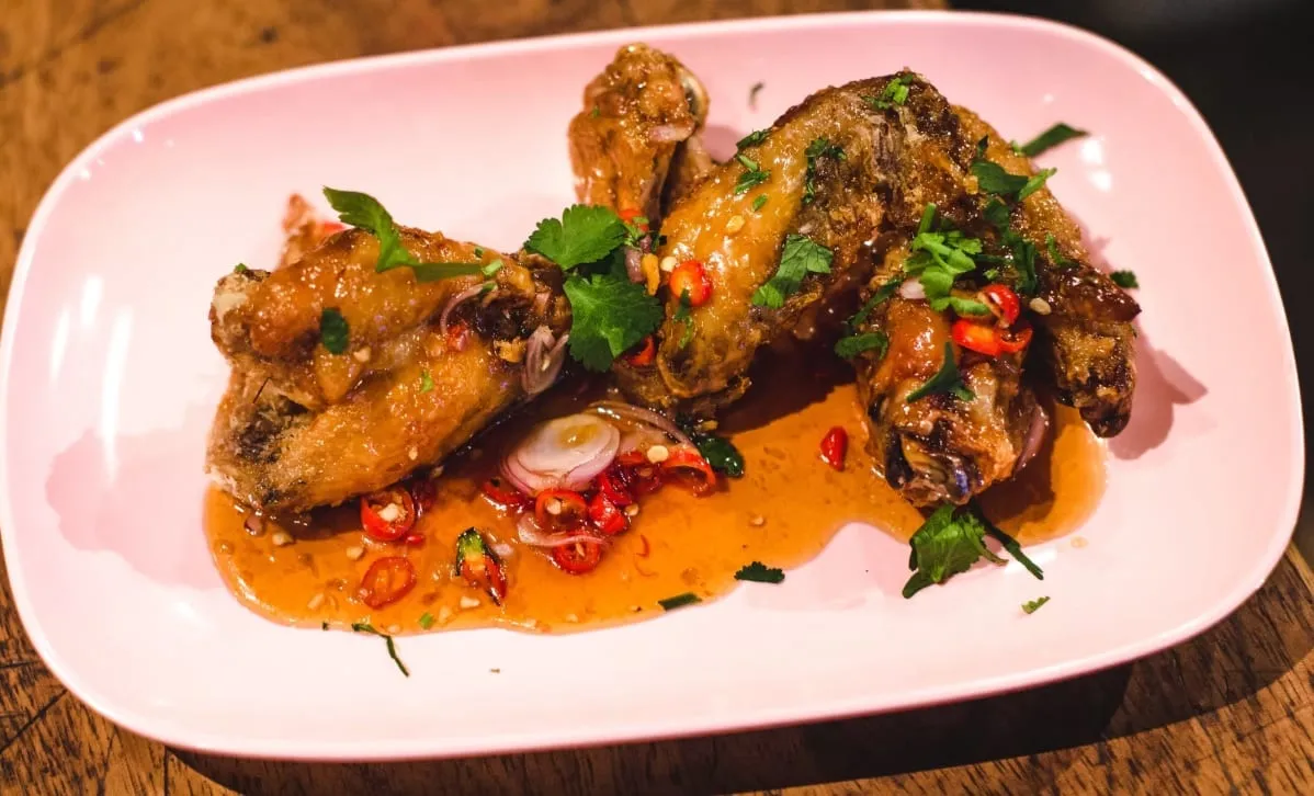 Glazed chicken wings on a pink oval plate, scattered with coriander, sliced red chilli and shallot in a pool of orange fish sauce dressing