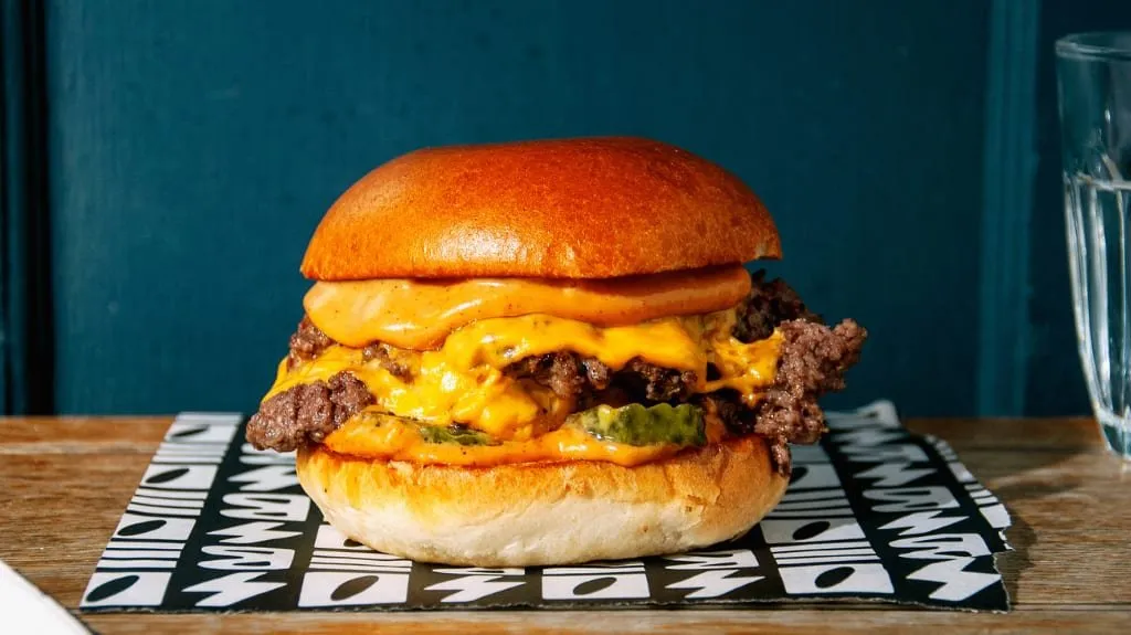 A SoBe Burger double smash burger on black-and-white patterned paper: two crisp, ragged-edged patties under melted cheese, pickles and an orange sauce in a glossy brioche bun