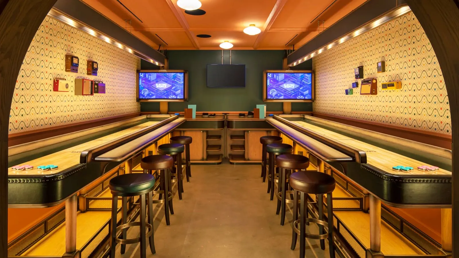 Two shuffleboard lanes facing each other in a room with patterned wallpaper, vintage radios on the walls, leather bar stools and a screen at the end of each lane