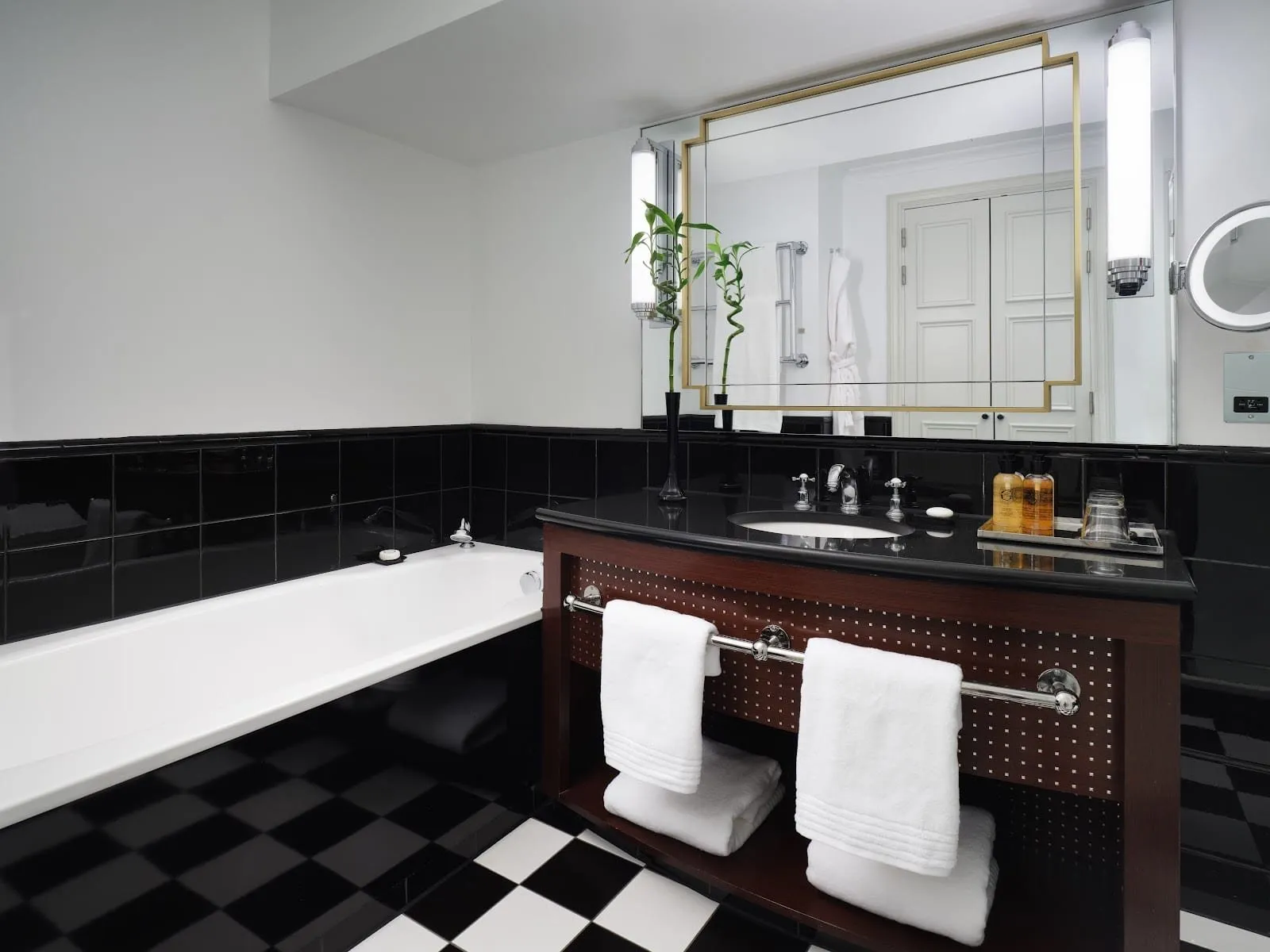A bathroom at Sofitel London St James with black wall tiles, a black-and-white chequerboard floor, a bath, and a basin set in a black counter under a gold-framed mirror